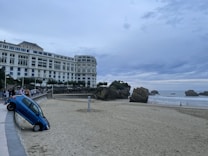 A seaside landscape with a large, elegant white building along a beach promenade. In the foreground, a blue car is precariously positioned halfway off a low wall leading down to the sandy beach. To the right, rock formations are present along the shore, and the ocean stretches into the distance under a cloudy sky. Several people are walking along the promenade and the beach.