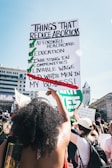 People are gathered in a public space, participating in a demonstration. A large sign is prominently held by an individual, listing points about reducing abortions, highlighting affordable healthcare, education, and other social investments, while crossing out old white men's involvement in personal matters. The setting appears to be urban, with detailed architecture visible in the background.