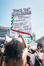 People are gathered in a public space, participating in a demonstration. A large sign is prominently held by an individual, listing points about reducing abortions, highlighting affordable healthcare, education, and other social investments, while crossing out old white men's involvement in personal matters. The setting appears to be urban, with detailed architecture visible in the background.