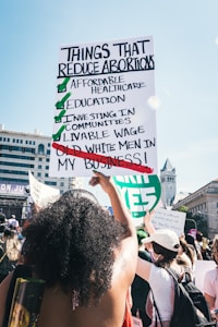 People are gathered in a public space, participating in a demonstration. A large sign is prominently held by an individual, listing points about reducing abortions, highlighting affordable healthcare, education, and other social investments, while crossing out old white men's involvement in personal matters. The setting appears to be urban, with detailed architecture visible in the background.