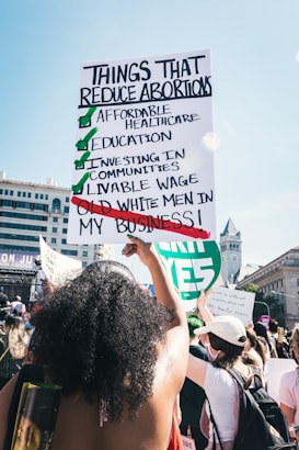 People are gathered in a public space, participating in a demonstration. A large sign is prominently held by an individual, listing points about reducing abortions, highlighting affordable healthcare, education, and other social investments, while crossing out old white men's involvement in personal matters. The setting appears to be urban, with detailed architecture visible in the background.