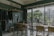 A cozy cafe interior with wooden tables and chairs positioned near large glass windows that overlook a green, tree-filled area outside. On one wall, there are clothes hanging on a rack, indicating a potential retail section. The lighting is soft, and small plants in glass bottles decorate the tables and window sills.