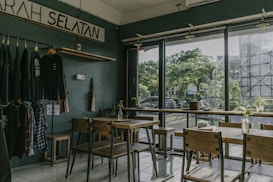 A cozy cafe interior with wooden tables and chairs positioned near large glass windows that overlook a green, tree-filled area outside. On one wall, there are clothes hanging on a rack, indicating a potential retail section. The lighting is soft, and small plants in glass bottles decorate the tables and window sills.