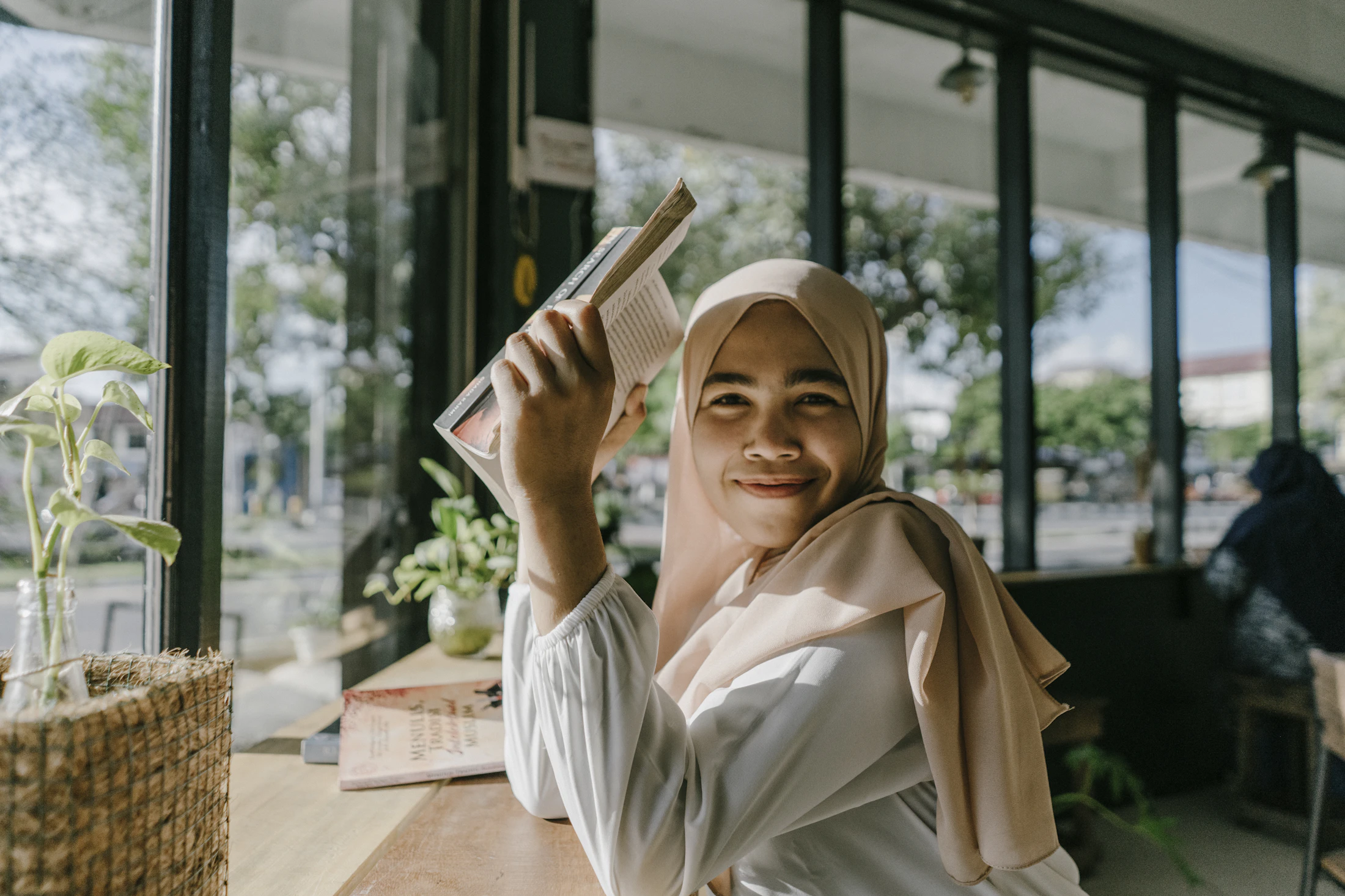 A warm, smiling teacher in a hijab holding a book by a window