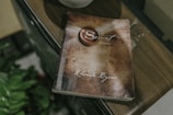 A book with a worn cover titled 'The Secret' by Rhonda Byrne is lying on a wooden surface. There are some green leaves partially visible at the bottom left corner, suggesting the presence of a plant nearby. The lighting and tone suggest a serene, contemplative atmosphere.
