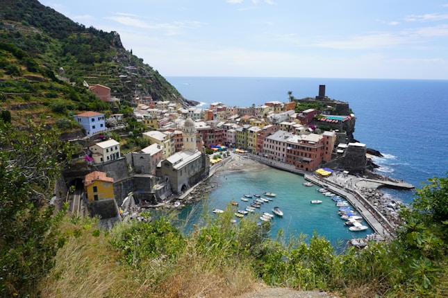 A coastal village with colorful buildings surrounding a small harbor. Boats are anchored in the clear turquoise water. The village is set against a backdrop of lush green hills and the expansive blue sea.