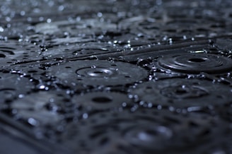 A close-up view of metal gears and cogs covered in droplets of water, with a focus on the textures and patterns of the machinery. The surface appears wet and shiny, enhancing the industrial feel.