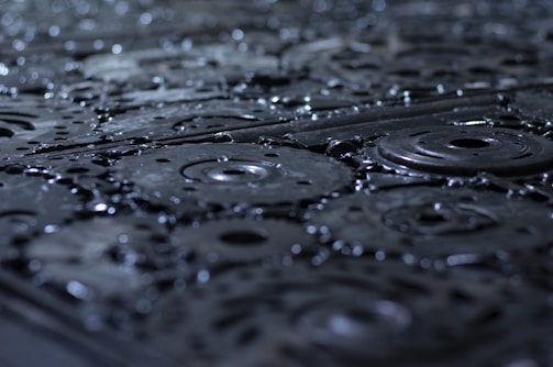 A close-up view of metal gears and cogs covered in droplets of water, with a focus on the textures and patterns of the machinery. The surface appears wet and shiny, enhancing the industrial feel.