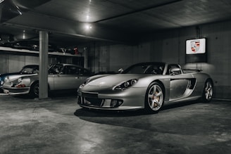 A dimly lit garage houses two sleek silver sports cars, with one prominently displayed in the foreground and the other slightly obscured behind a concrete column. The foreground car is a modern, aerodynamic convertible, and the background seems to feature a classic model. The walls are plain concrete, and a Porsche emblem is mounted on the wall.