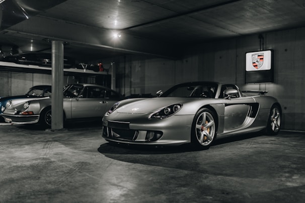A dimly lit garage houses two sleek silver sports cars, with one prominently displayed in the foreground and the other slightly obscured behind a concrete column. The foreground car is a modern, aerodynamic convertible, and the background seems to feature a classic model. The walls are plain concrete, and a Porsche emblem is mounted on the wall.