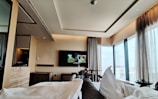 A modern hotel room with large floor-to-ceiling windows providing a view of the cityscape. The room features a sleek, contemporary design with neutral-toned walls and furnishings. A television is mounted on the wall opposite the bed, displaying a scene from a movie. Comfortable bedding and curtains add a cozy atmosphere.