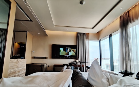 A modern hotel room with large floor-to-ceiling windows providing a view of the cityscape. The room features a sleek, contemporary design with neutral-toned walls and furnishings. A television is mounted on the wall opposite the bed, displaying a scene from a movie. Comfortable bedding and curtains add a cozy atmosphere.