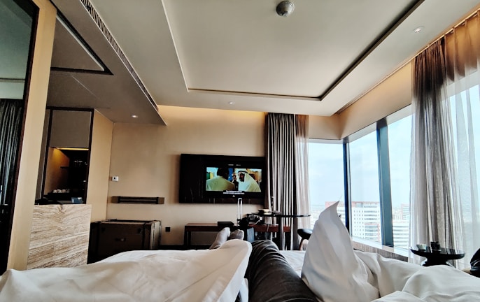A modern hotel room with large floor-to-ceiling windows providing a view of the cityscape. The room features a sleek, contemporary design with neutral-toned walls and furnishings. A television is mounted on the wall opposite the bed, displaying a scene from a movie. Comfortable bedding and curtains add a cozy atmosphere.