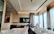 A modern hotel room with large floor-to-ceiling windows providing a view of the cityscape. The room features a sleek, contemporary design with neutral-toned walls and furnishings. A television is mounted on the wall opposite the bed, displaying a scene from a movie. Comfortable bedding and curtains add a cozy atmosphere.