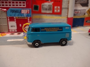 A blue toy van with the text 'MATCHBOX INTERNATIONAL EXPRESS' displayed on its side is placed on a surface. The background features a model setup including signs such as 'FORMULA DRIFT' and a gas station scene. There are various colorful elements and signs in the backdrop indicating a miniature diorama setup.