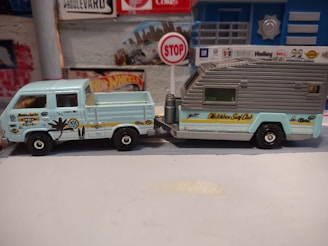 A small, detailed model of a vintage Volkswagen pickup truck attached to a camper trailer is displayed. The pickup is light blue with various surf-themed decals, including a palm tree and the words 'Locals Only.' The trailer has a grey roof with the words 'Matchbox Surf Club' written on the side in yellow. The background includes a wall with a Hot Wheels sign and a small stop sign, creating a miniature diorama setting.