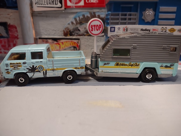 A small, detailed model of a vintage Volkswagen pickup truck attached to a camper trailer is displayed. The pickup is light blue with various surf-themed decals, including a palm tree and the words 'Locals Only.' The trailer has a grey roof with the words 'Matchbox Surf Club' written on the side in yellow. The background includes a wall with a Hot Wheels sign and a small stop sign, creating a miniature diorama setting.