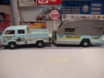 A small, detailed model of a vintage Volkswagen pickup truck attached to a camper trailer is displayed. The pickup is light blue with various surf-themed decals, including a palm tree and the words 'Locals Only.' The trailer has a grey roof with the words 'Matchbox Surf Club' written on the side in yellow. The background includes a wall with a Hot Wheels sign and a small stop sign, creating a miniature diorama setting.