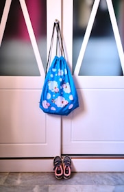 A drawstring bag with a cheerful design featuring clouds and cartoon characters hangs on a white paneled door. Below the bag, a pair of children's sneakers is neatly placed on a tiled floor.