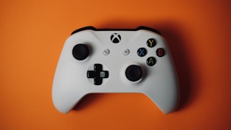A white gaming controller with an asymmetrical design, featuring two thumbsticks, a directional pad, and four colorful buttons labeled Y, B, A, and X, resting on an orange background.