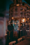 A vintage-style hotel building with the sign 'Hotel Carlton' dominates the scene, reflecting urban life in a city. The image captures the interplay of light and reflections in a cafe or restaurant setting with people visible through glass, contributing to a layered and dynamic composition.
