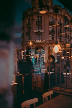 A vintage-style hotel building with the sign 'Hotel Carlton' dominates the scene, reflecting urban life in a city. The image captures the interplay of light and reflections in a cafe or restaurant setting with people visible through glass, contributing to a layered and dynamic composition.