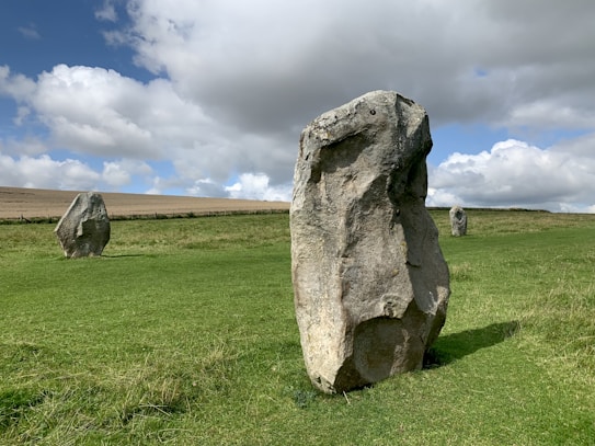 Large standing stones are situated in a grassy landscape under a partly cloudy sky. The stones are weathered and irregularly shaped, likely part of an ancient site or formation. The field extends into the distance bordered by a simple wooden fence.