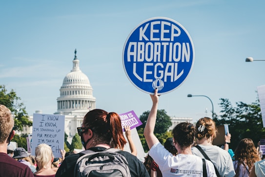 A group of people gather near a prominent building, holding various protest signs advocating for keeping abortion legal. The atmosphere suggests a demonstration or rally with individuals of diverse backgrounds actively participating.