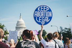 A group of people gather near a prominent building, holding various protest signs advocating for keeping abortion legal. The atmosphere suggests a demonstration or rally with individuals of diverse backgrounds actively participating.