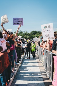 A large group of people is gathered for a protest. Many individuals are holding signs with messages advocating for women's rights and reproductive freedom. The crowd is lined up on either side of a pathway, with barriers separating them from the path where a few individuals are walking. The sky is clear and it appears to be a sunny day.