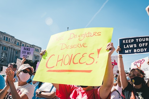 A group of people participating in a protest, holding various signs advocating for women's rights and bodily autonomy. The central sign, written in bold on bright yellow paper, says 'Our Daughters Deserve Choices.' Other visible signs include messages like 'Bans Off My Body' and 'Keep Your Laws Off My Body.' Many participants are wearing masks, and it appears to be daytime with a clear blue sky.