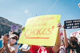 A group of people participating in a protest, holding various signs advocating for women's rights and bodily autonomy. The central sign, written in bold on bright yellow paper, says 'Our Daughters Deserve Choices.' Other visible signs include messages like 'Bans Off My Body' and 'Keep Your Laws Off My Body.' Many participants are wearing masks, and it appears to be daytime with a clear blue sky.