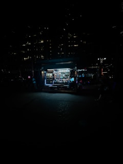 A dimly lit street scene featuring a small, illuminated kiosk or newsstand. The kiosk is filled with various products and stands out against the dark surroundings. The cityscape is barely visible in the background, with a few lights from nearby buildings and street lamps providing minimal illumination.