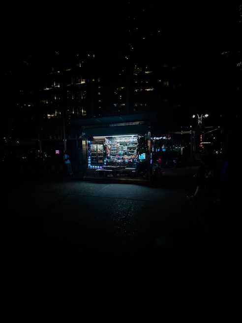 A dimly lit street scene featuring a small, illuminated kiosk or newsstand. The kiosk is filled with various products and stands out against the dark surroundings. The cityscape is barely visible in the background, with a few lights from nearby buildings and street lamps providing minimal illumination.