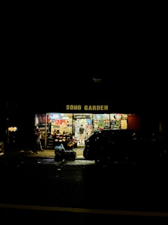 A small convenience store named 'SOHO GARDEN' is brightly lit, standing out against the dark surroundings at night. The storefront displays a variety of items, including fruits, flowers, and numerous signs with prices. There are people seen near the entrance, and a parked car is partially visible in front.