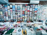 A wide selection of magazines and publications neatly displayed on a newsstand. Various magazine titles are stacked in piles on shelves, organized in rows. The covers feature diverse topics such as fashion, lifestyle, health, and sports with bright and colorful covers, featuring bold typography and striking images.