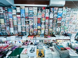 A wide selection of magazines and publications neatly displayed on a newsstand. Various magazine titles are stacked in piles on shelves, organized in rows. The covers feature diverse topics such as fashion, lifestyle, health, and sports with bright and colorful covers, featuring bold typography and striking images.