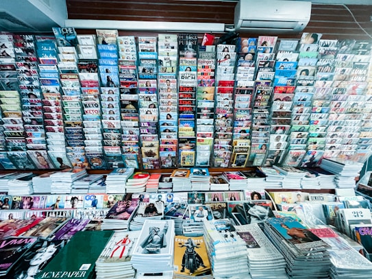 A wide selection of magazines and publications neatly displayed on a newsstand. Various magazine titles are stacked in piles on shelves, organized in rows. The covers feature diverse topics such as fashion, lifestyle, health, and sports with bright and colorful covers, featuring bold typography and striking images.