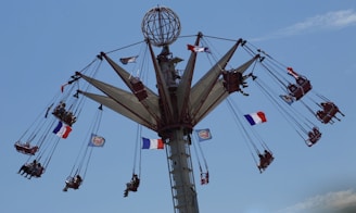 A high swing ride at an amusement park with several seats attached to long chains, spinning around a central column. The riders appear to be having fun as they fly through the air, and several French flags are visible, fluttering in the wind. The sky is clear and blue.