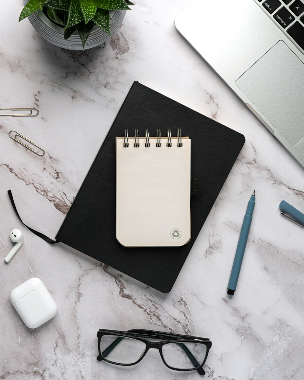 a desk with a notebook, pen, eyeglasses and a laptop