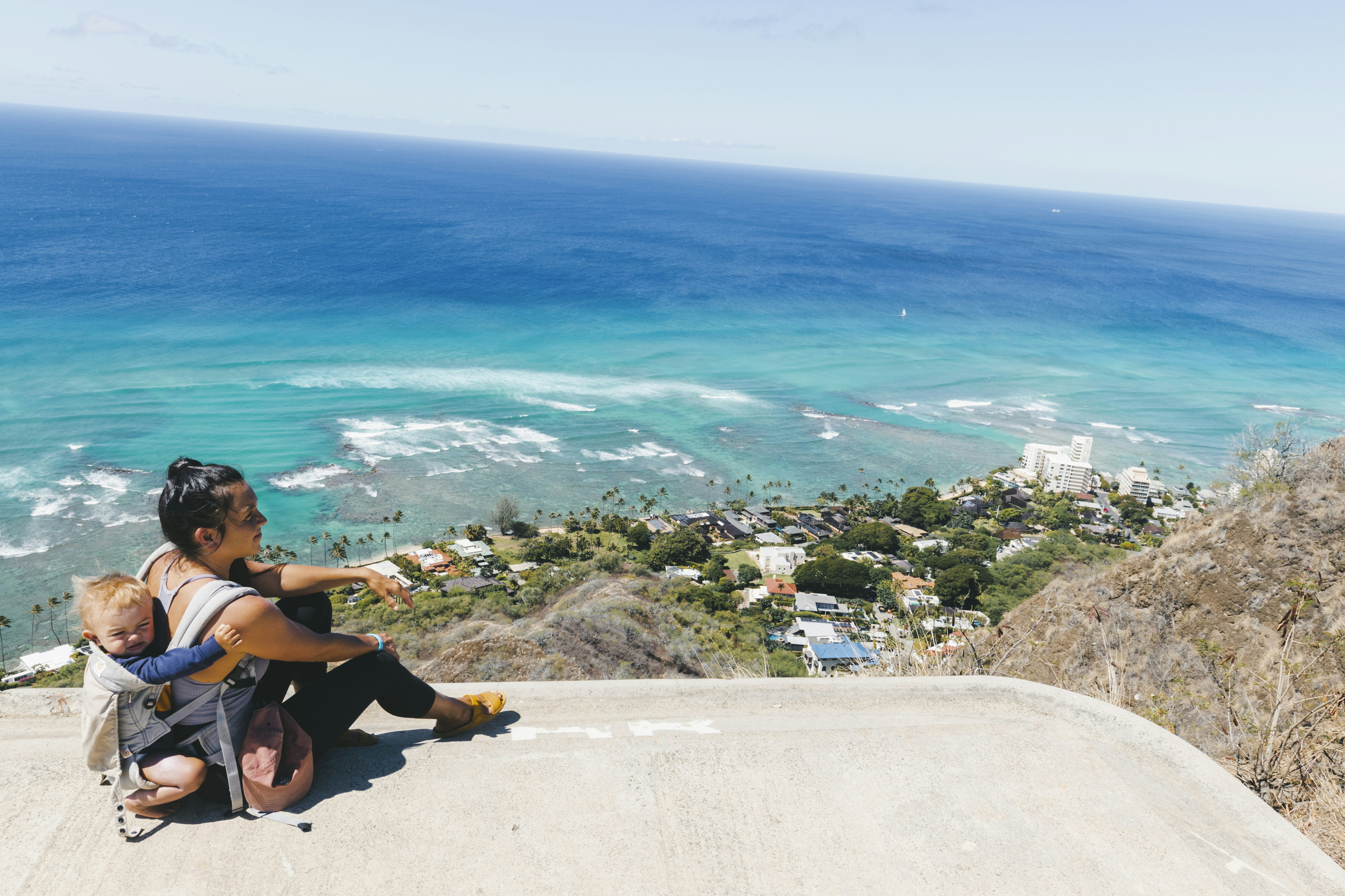 a woman and child sitting on a ledge overlooking the ocean, resting after a hard hike in hawaii
