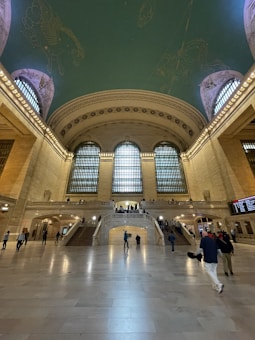 A large, grand interior space with a high, ornate ceiling decorated with intricate designs. The room is filled with natural light coming through oversized arched windows. People are walking across a spacious marble floor and ascending wide staircases. The architectural design is classic and opulent, with detailed stonework and elegant railings.