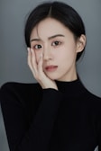 A person with dark hair poses with their hand gently touching the side of their face, wearing a black ribbed turtleneck sweater. The background is a plain, muted gray, adding to the minimalist and elegant appearance.