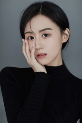 A person with dark hair poses with their hand gently touching the side of their face, wearing a black ribbed turtleneck sweater. The background is a plain, muted gray, adding to the minimalist and elegant appearance.