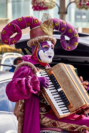 A person dressed in a colorful and elaborate jester costume, featuring an intricate headdress with large curved purple horns and a mask with painted facial features. The outfit is embellished with vibrant details and textures. The individual is playing an accordion against the backdrop of a street scene.