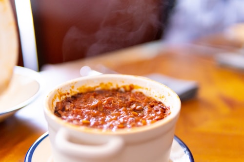 A white ceramic bowl filled with a steaming hot dish, possibly a baked or stewed meal, with a browned or caramelized surface. The bowl is placed on a saucer with a blurred warm-colored background, suggesting a cozy dining setting.