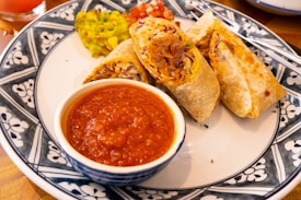 A plate containing a few pieces of burrito or wrap, filled with meat and vegetables, is served with a bowl of red sauce. Garnishes of guacamole and diced tomatoes are also present on the plate. The dish is presented on a beautifully patterned plate with a blue and white design.