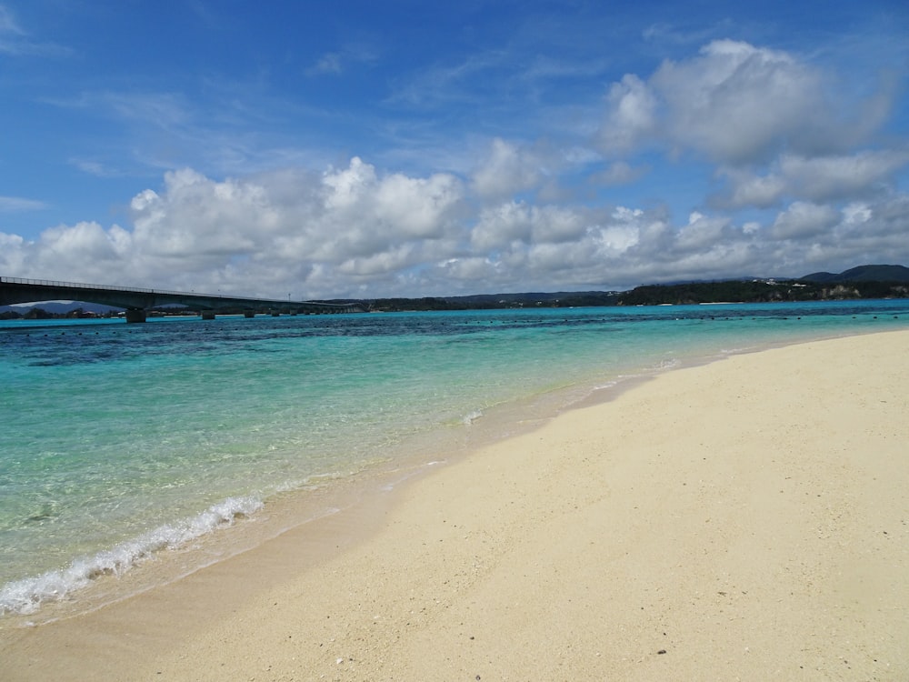 a sandy beach with clear blue water and a bridge in the distance