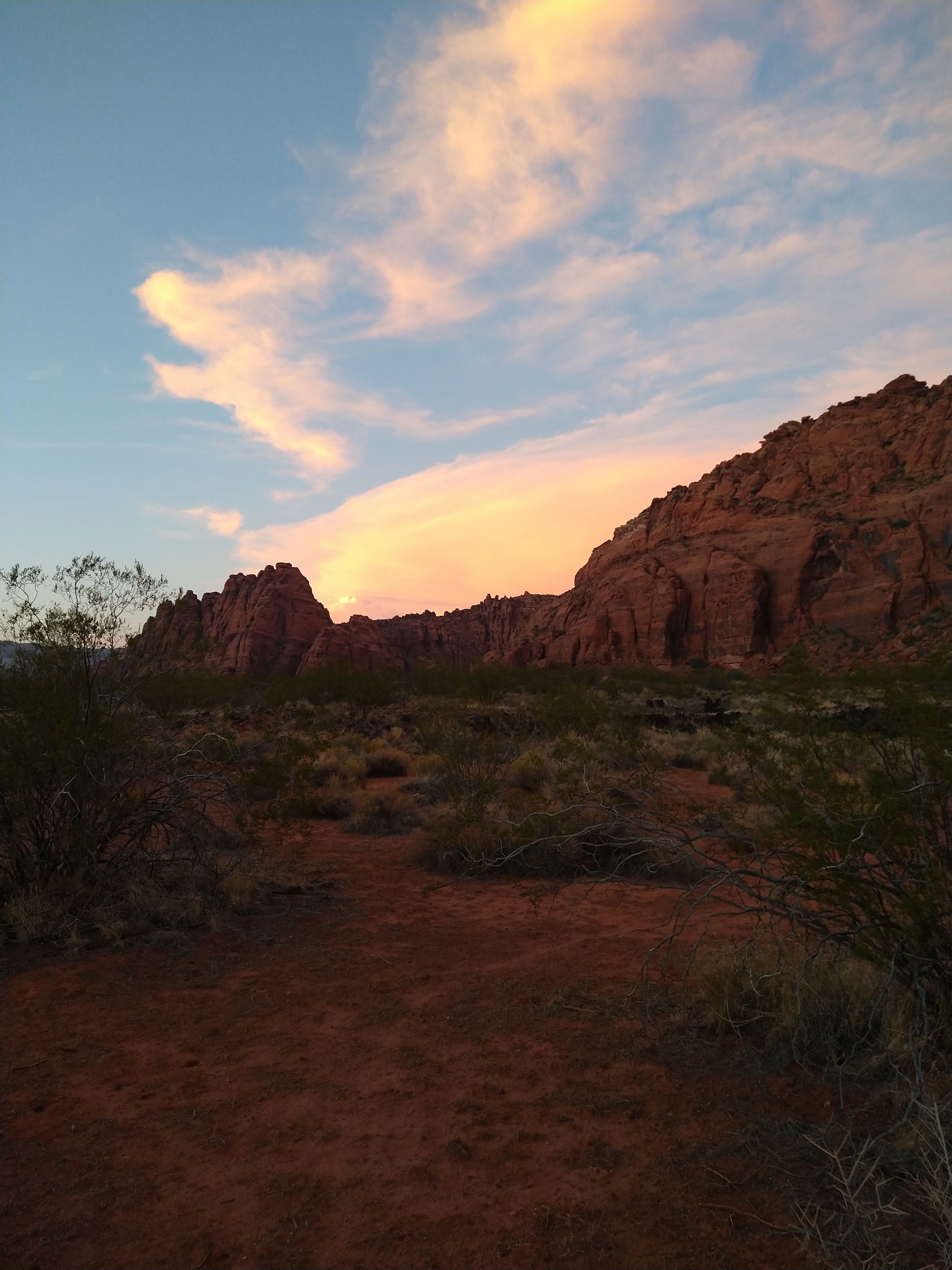 A wide-open desert landscape under a vibrant sunset sky, with red rock formations casting long shadows.
