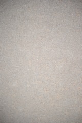 A close-up view of a textured surface that appears to be a concrete or cement wall. The surface is uniform in color with small specks and imperfections throughout, giving it a rough, gritty appearance.